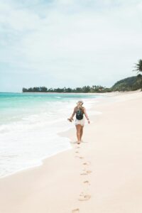 A woman with a backpack walks barefoot along a pristine tropical beach, leaving footprints in the sand.