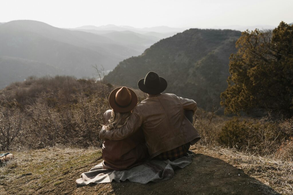 Back view anonymous couple in trendy leather jackets and hats embracing and admiring scenic hilly terrain while sitting on hilltop